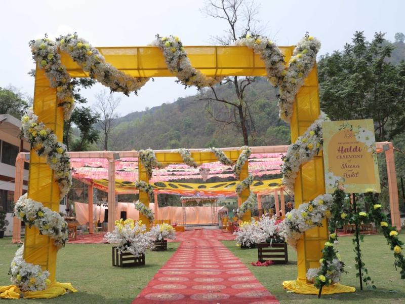 haldi ceremony entrance with a vibrant yellow archway wrapped in white floral garlands, leading to a beautifully decorated open air venue with drapes