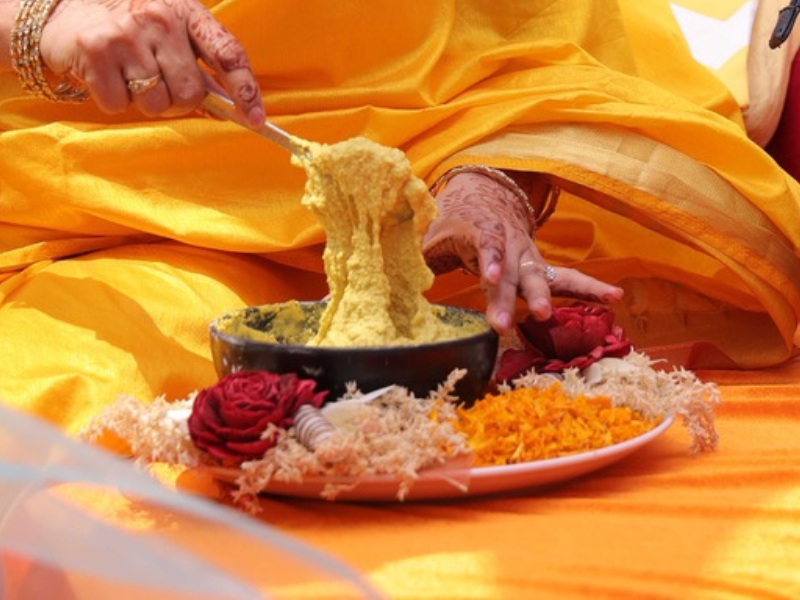 woman dressed in a yellow saree preparing a turmeric paste for the haldi ceremony rituals