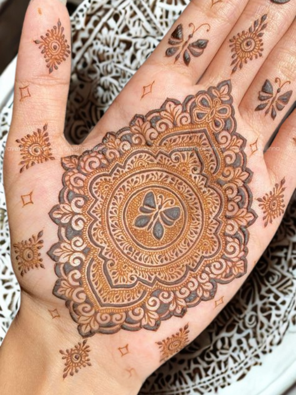 Intricate Mehndi design on the palm featuring a large central mandala with a butterfly motif, surrounded by fine floral and paisley patterns, with additional small butterfly and geometric accents on the fingers and wrist.