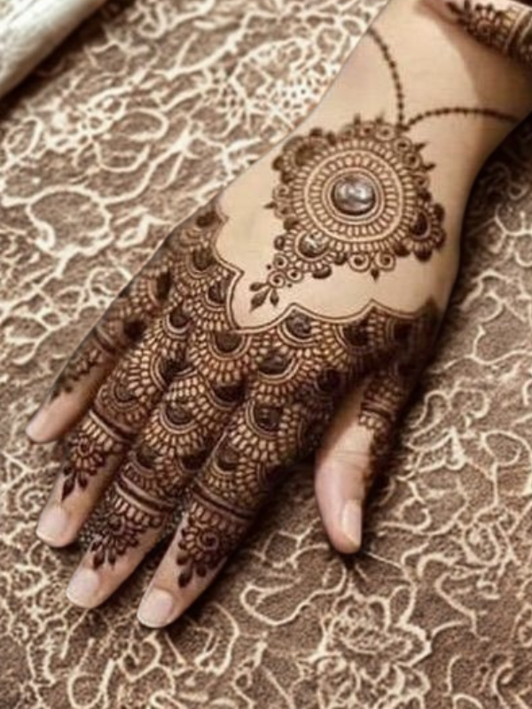 Intricate bridal Mehndi design on the back of the hand featuring a large circular mandala pattern connected to the wrist with dotted chains, and symmetrical scalloped motifs extending over the fingers